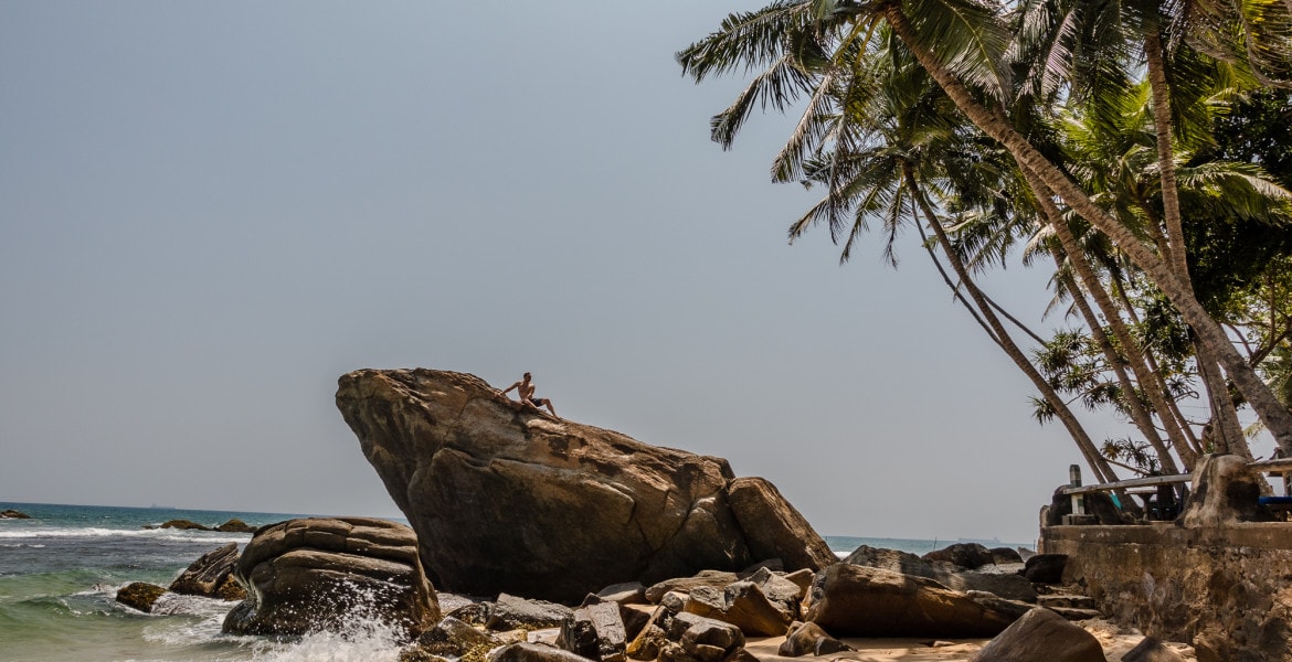 Man sitting on a gigantic rock on the beach with palmtrees lining the beach of Dalawella near Unawatuna in Sri Lanka