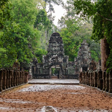 Sandy pathway with rain puddles lined with statues and green luscious trees leading to the stone entrance gate of the temple of Preah Khan inside the Angkor Wat Complex in Cambodia.